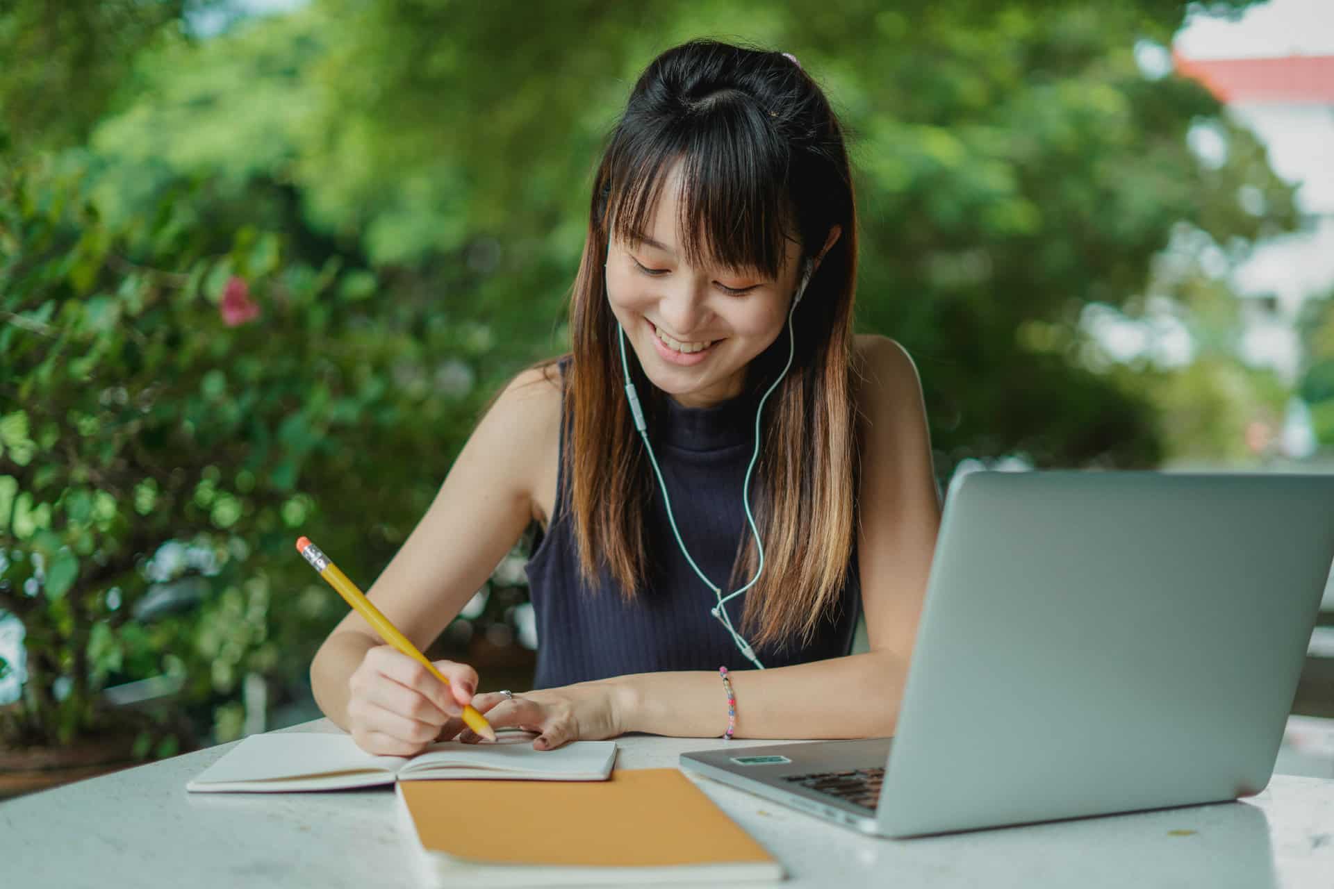 A student writing in a notebook with a laptop in front of her.
