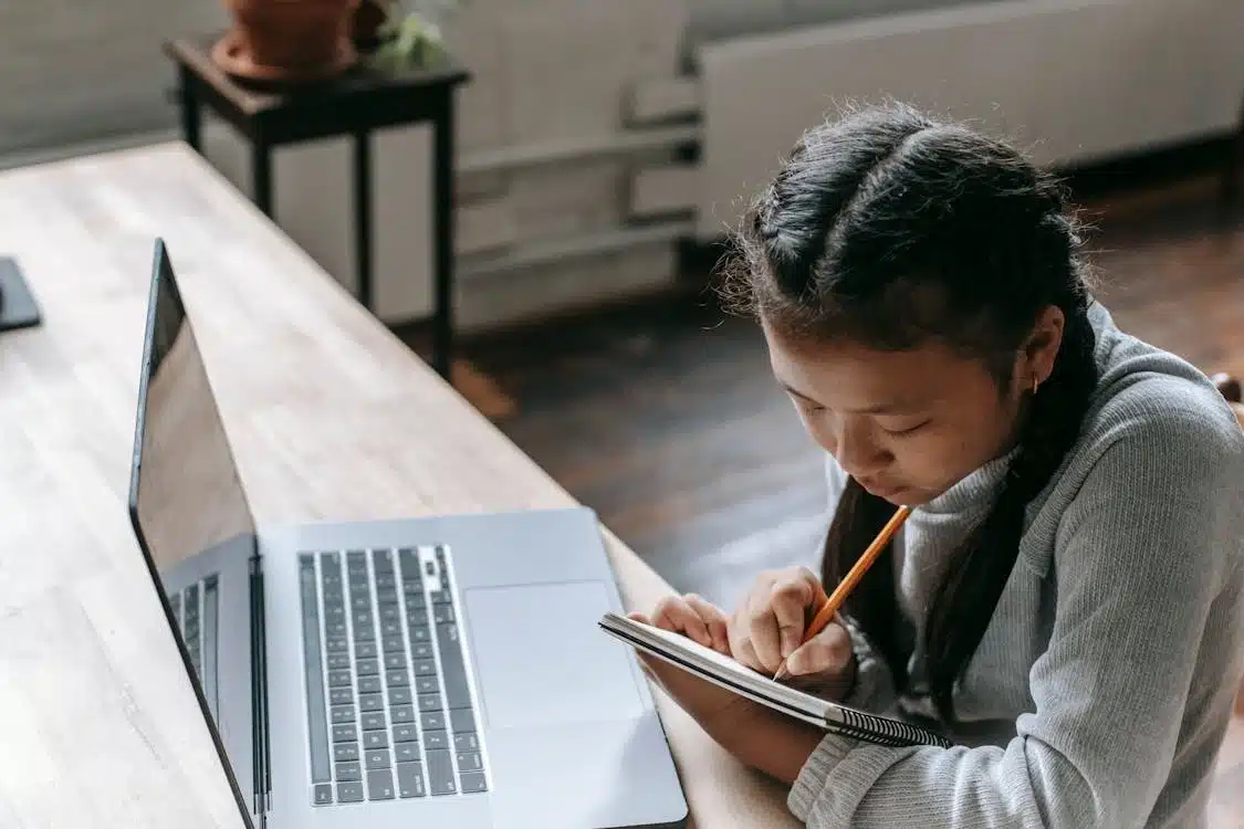 A student writing in her notebook with a laptop in front of her.